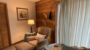 Una habitación acogedora con una silla de mimbre, una mesita con una lámpara, un cuadro enmarcado en la pared y una ventana con cortinas.