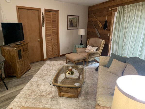 Una sala de estar con una televisión sobre un mueble de madera, una silla de mimbre, un sofá con cojines y una mesa de centro de vidrio con una vela.