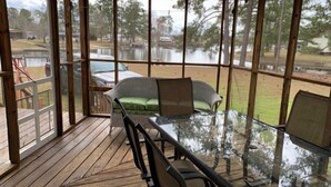 Un espacio al aire libre cubierto con una mesa y sillas de vidrio, un sofá de mimbre con cojines, y vistas a un lago y árboles circundantes.
