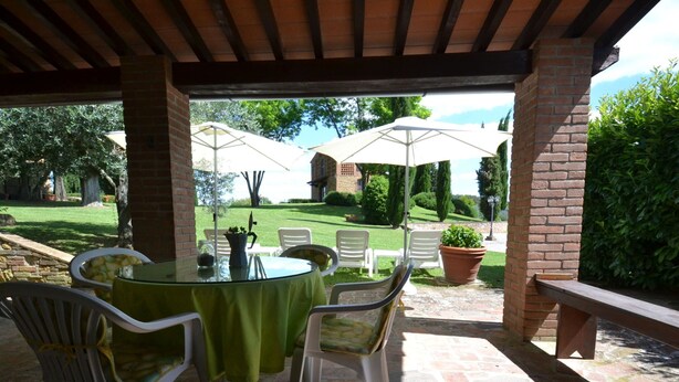 Un patio esterno con un tavolo rotondo e sedie, una colonna in mattoni e vista su un prato e un edificio sullo sfondo.