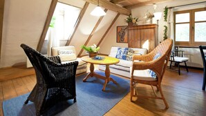 A cozy loft-style room with a high ceiling, exposed wooden beams, a round wooden table, wicker chairs, a sofa with patterned cushions, and a radiator. - Superb Holiday Home in Weissenburg near Forest (Weissenburg in Bayern)