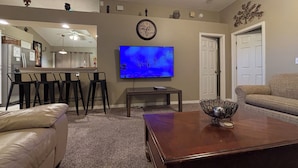 A living room with a flat-screen TV, a wooden coffee table, a sofa, and a kitchen area in the background.