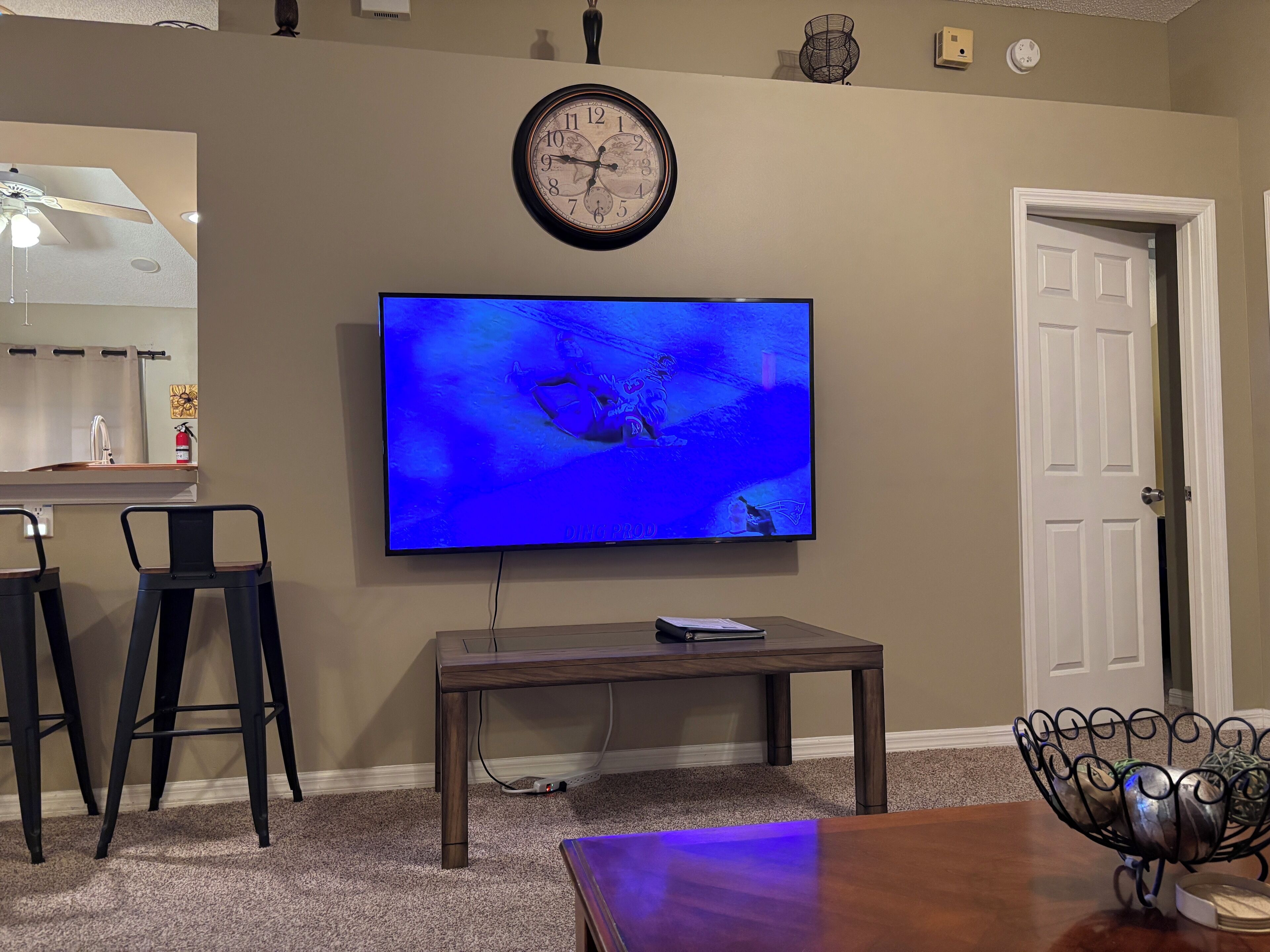 A living room with a flat-screen TV mounted on the wall, a wooden coffee table, two black bar stools, a dining table, and a decorative metal bowl on the floor.