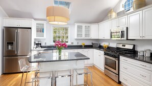 A modern kitchen with white cabinets, stainless steel appliances, a central island with bar stools, and a large window providing natural light. - Hingham Harbor Retreat ~ Sunsets ~ Family Gatherings ~ Trails at World’s End (Hingham,)