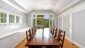 A long dining table with chairs, white cabinetry, and a view to an outdoor patio. - Hingham Harbor Retreat ~ Sunsets ~ Family Gatherings ~ Trails at World’s End (Hingham,)