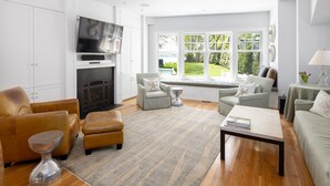 A modern living room with a fireplace, comfortable seating, and large windows offering a view of the outdoors. - Hingham Harbor Retreat ~ Sunsets ~ Family Gatherings ~ Trails at World’s End (Hingham,)