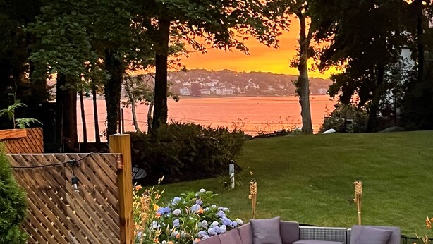 A patio with outdoor furniture, a wooden fence, and a view of a body of water during sunset. - Hingham Harbor Retreat ~ Sunsets ~ Family Gatherings ~ Trails at World’s End (Hingham,)