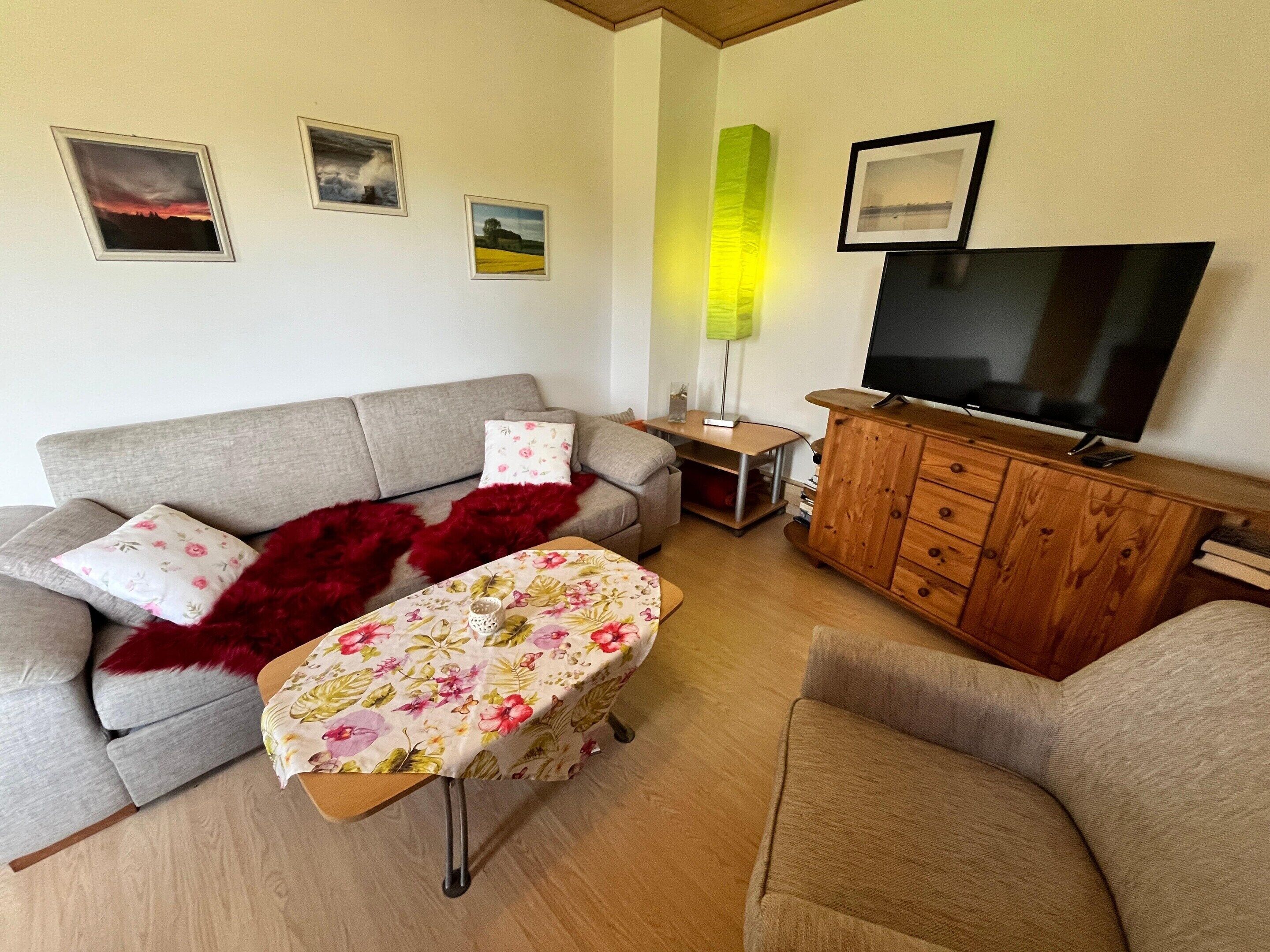 A living room with a sofa, a table with a floral tablecloth, a television on a wooden cabinet, and framed pictures on the wall.