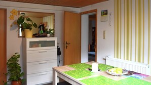 Ett rum med ett träpanelat tak, en vit byrå med lådor, en spegel, en krukväxt, ett bord med ett grönt bordsduk och ett element.