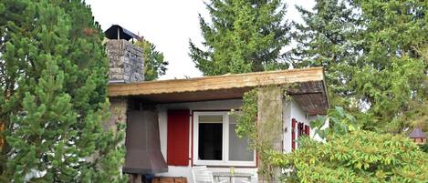 A rustic cabin with a wooden deck, a dining area with outdoor chairs, and a red door.