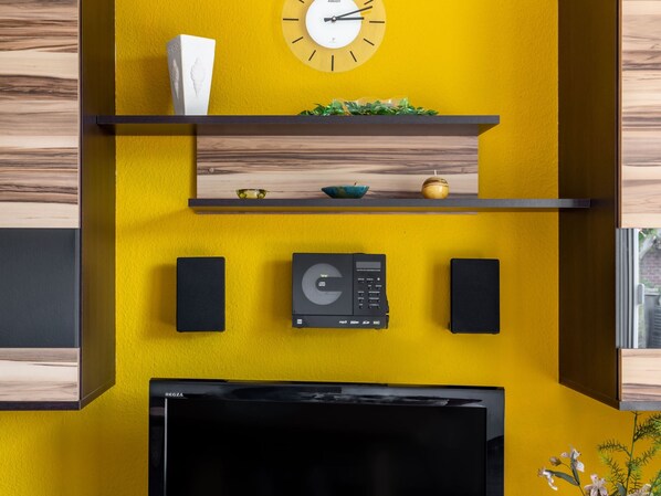 Ein modernes Wohnzimmer mit gelber Akzentwand, einer Uhr, einem Flachbildfernseher und eingebauten Lautsprechern.