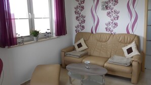 Un salon avec un canapé beige, deux coussins, une petite table ronde et une fenêtre ornée de rideaux violets.
