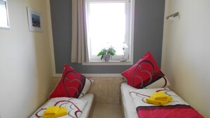 Une petite pièce étroite, avec deux lits simples, chacun agrémenté de draps rouges et blancs et de coussins assortis. Une fenêtre offre une vue sur la verdure environnante, et une petite table est placée près de la fenêtre, sur laquelle se trouvent une lampe et un pot de fleurs.