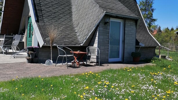 Een huis met een steil dak, een stenen schoorsteen en een klein terras met buitenmeubilair.