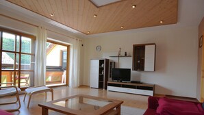 Ett rymligt vardagsrum med ett träpanelat tak, ett soffbord i glas, en röd soffa, en tv på ett träställ och en skjutdörr i glas som leder ut till ett utomhusområde.