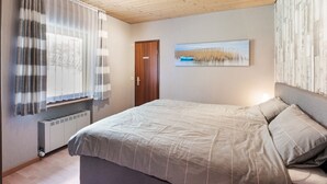 Ein Schlafzimmer mit Holzboden, einem Bett mit grauer Bettdecke, einem Fenster mit durchscheinenden Vorhängen, einem Heizkörper und einer Tür.