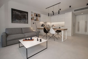 Een moderne woonkamer met een grijze bank, een witte salontafel en zwarte stoelen. Er is een keuken met witte kasten en een eettafel.