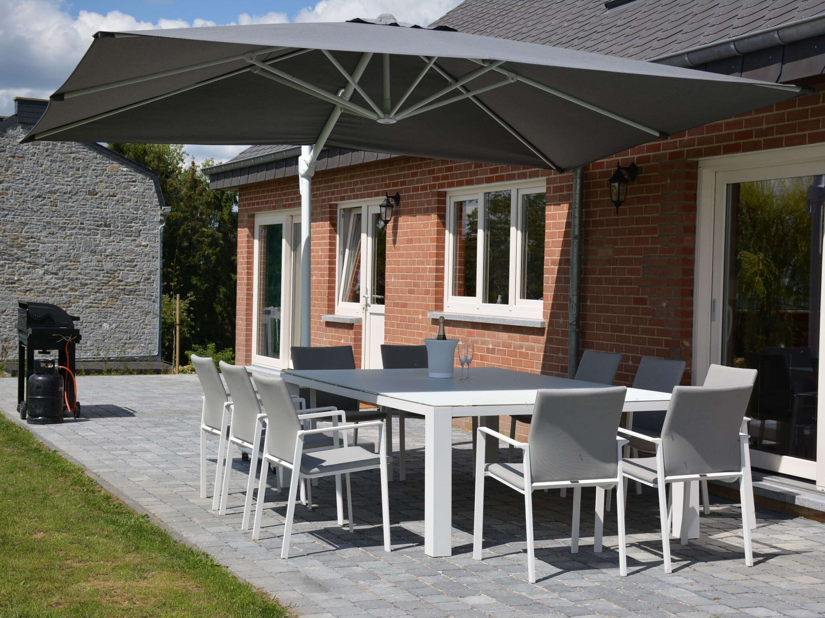An outdoor patio with a white table and chairs, a large umbrella, and a barbecue grill.