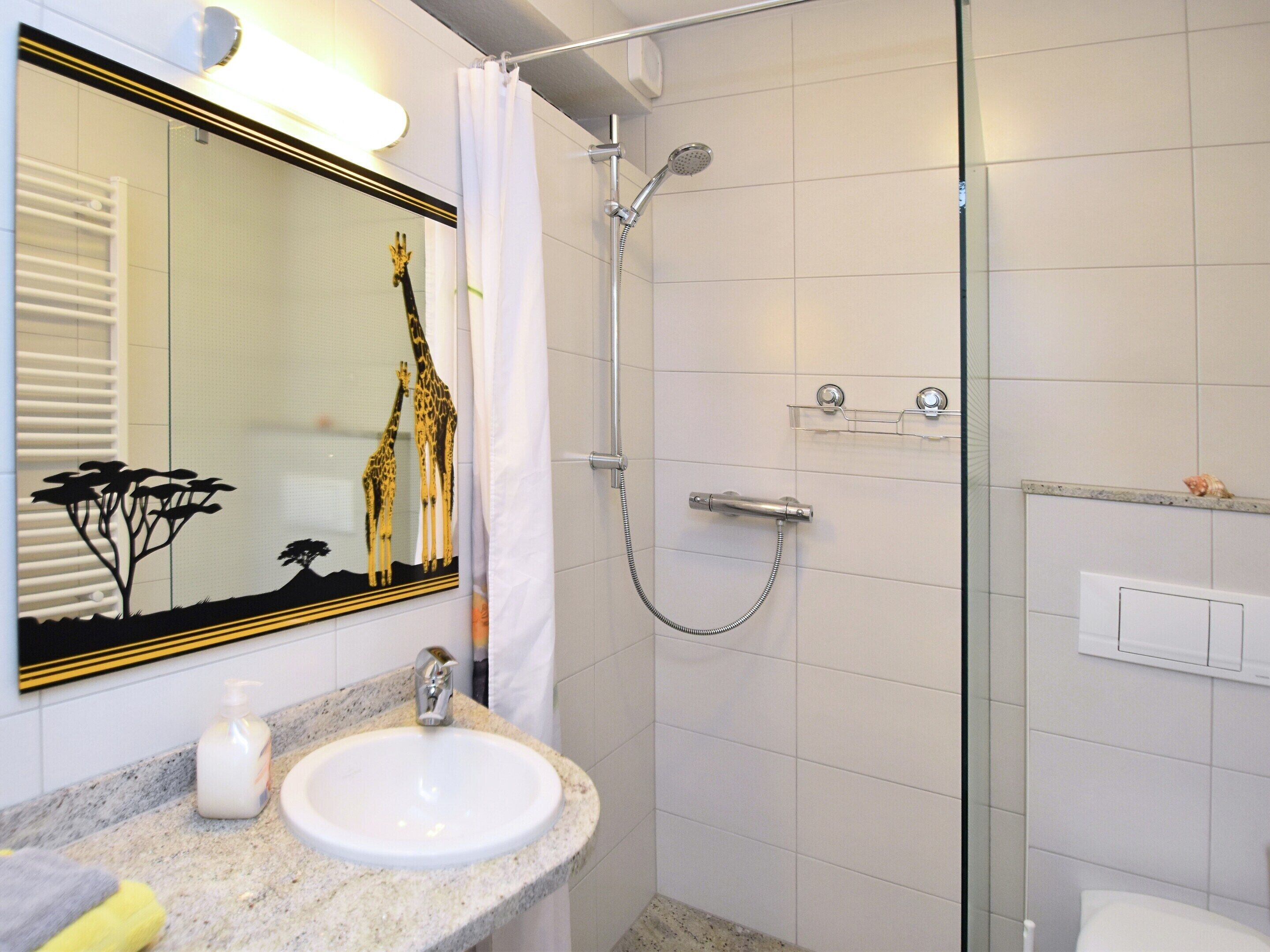A bathroom with a sink, mirror, shower, and a giraffe decoration on the wall.