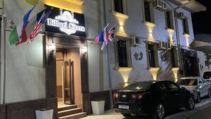 L’entrée d’un hôtel, avec une enseigne indiquant « Billiardi & Siera », des drapeaux et des voitures garées.