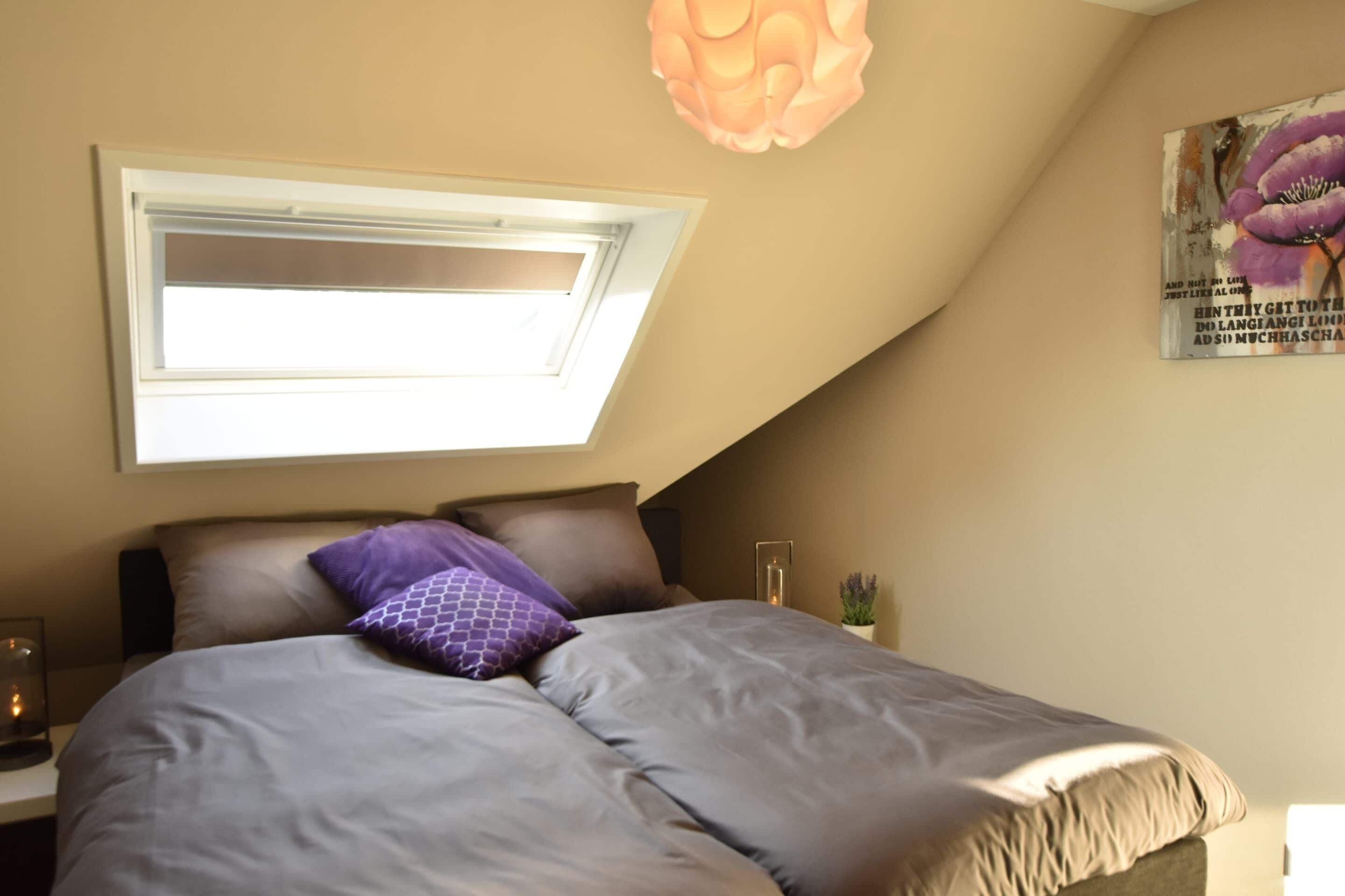 A bedroom with a skylight, a bed with grey bedding, a purple pillow, and a wall-mounted light fixture.