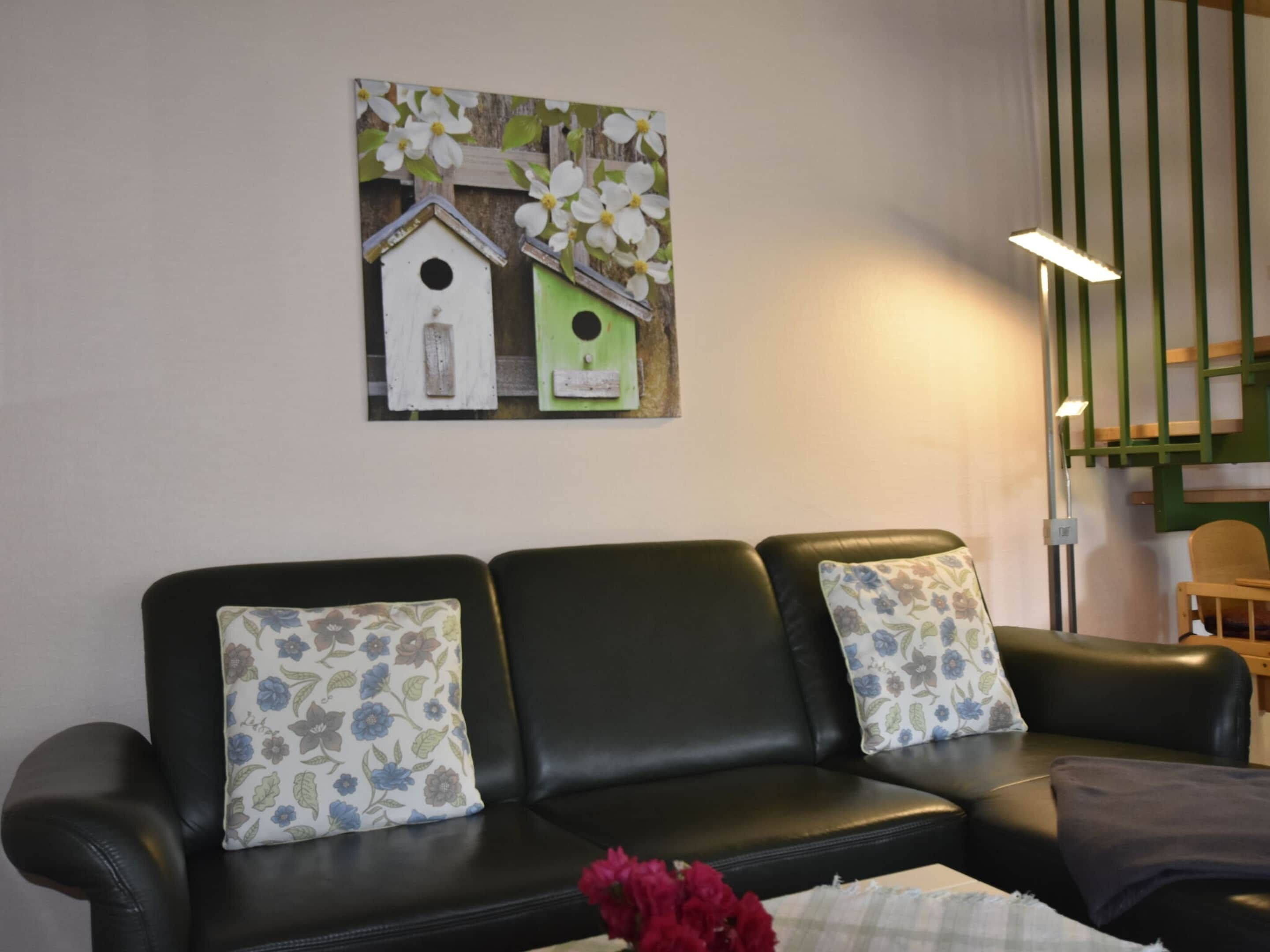 A black leather sofa with floral-patterned cushions, a wall-mounted artwork of birdhouses, and a small red flower arrangement.