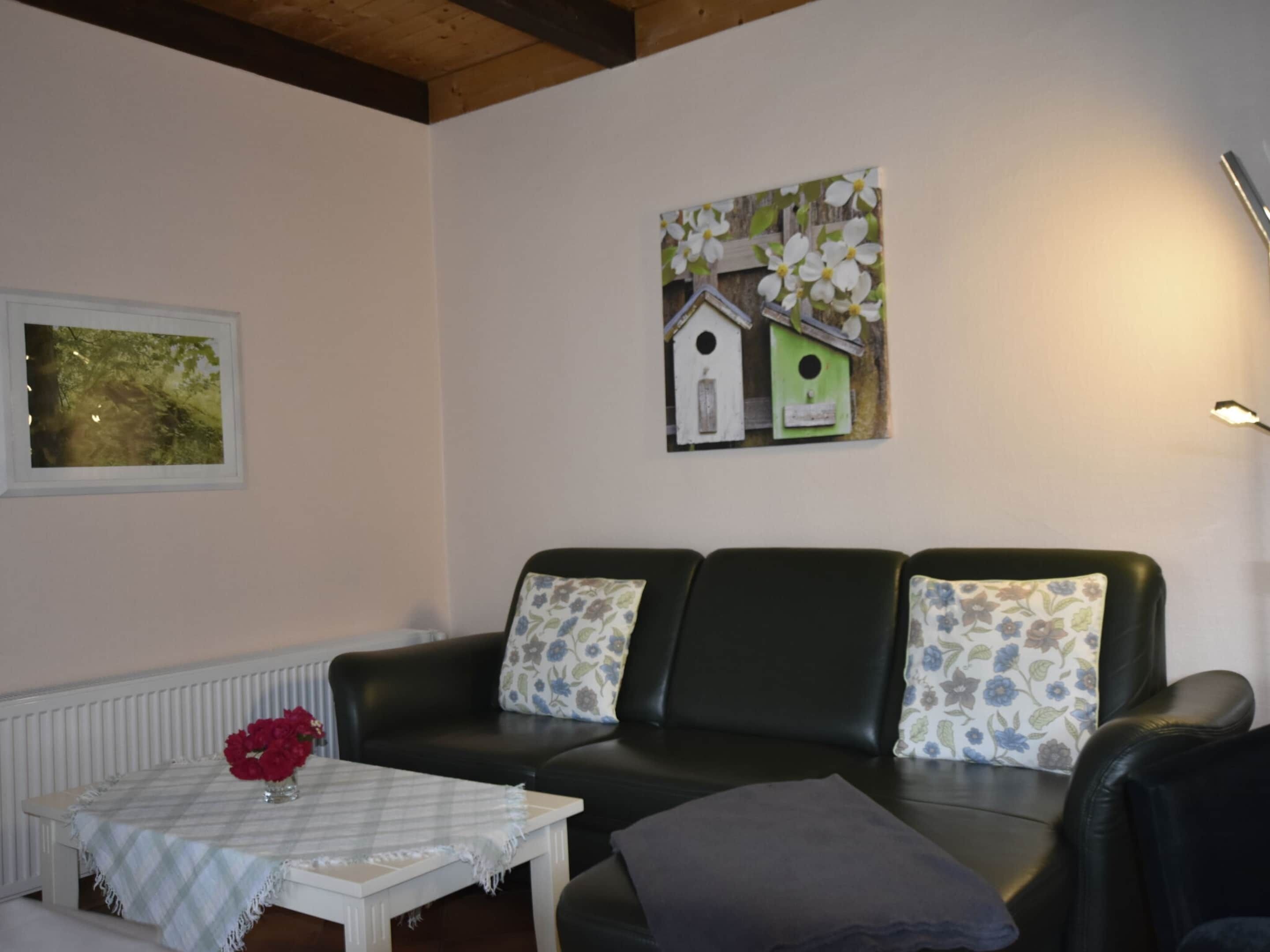 A living room with a black leather sofa, a white table with a plaid tablecloth, and a vase of red flowers. There is a framed picture of birds on the wall.