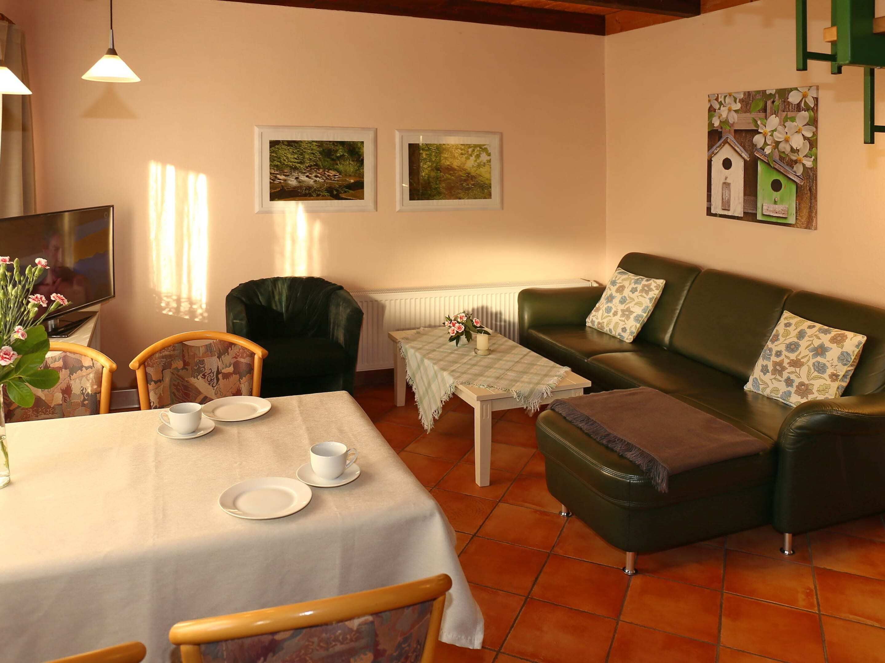Ein gemütliches Wohnzimmer mit Esstisch, einer Couch und zwei Sesseln. An der Wand hängen Bilder in Rahmen und darunter befindet sich eine Heizung.