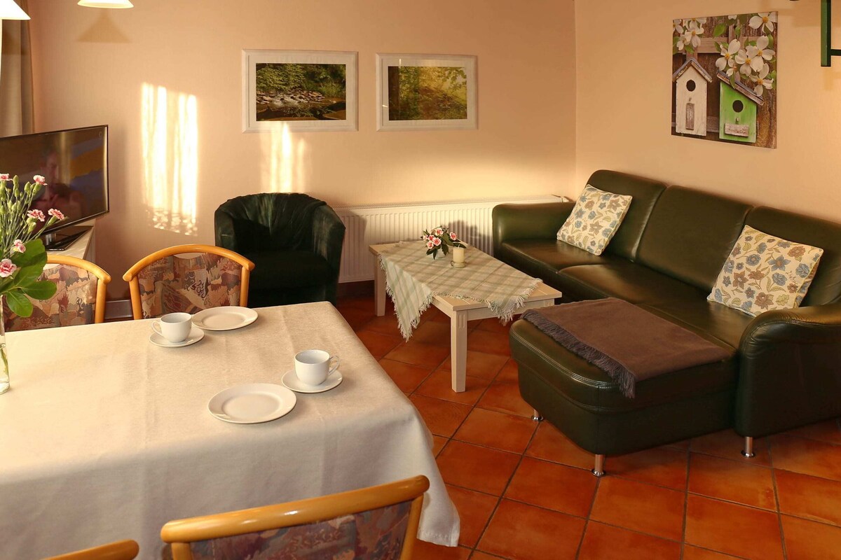 A cozy living room with a dining table, a sofa, and two armchairs. There are framed pictures on the wall and a radiator below.