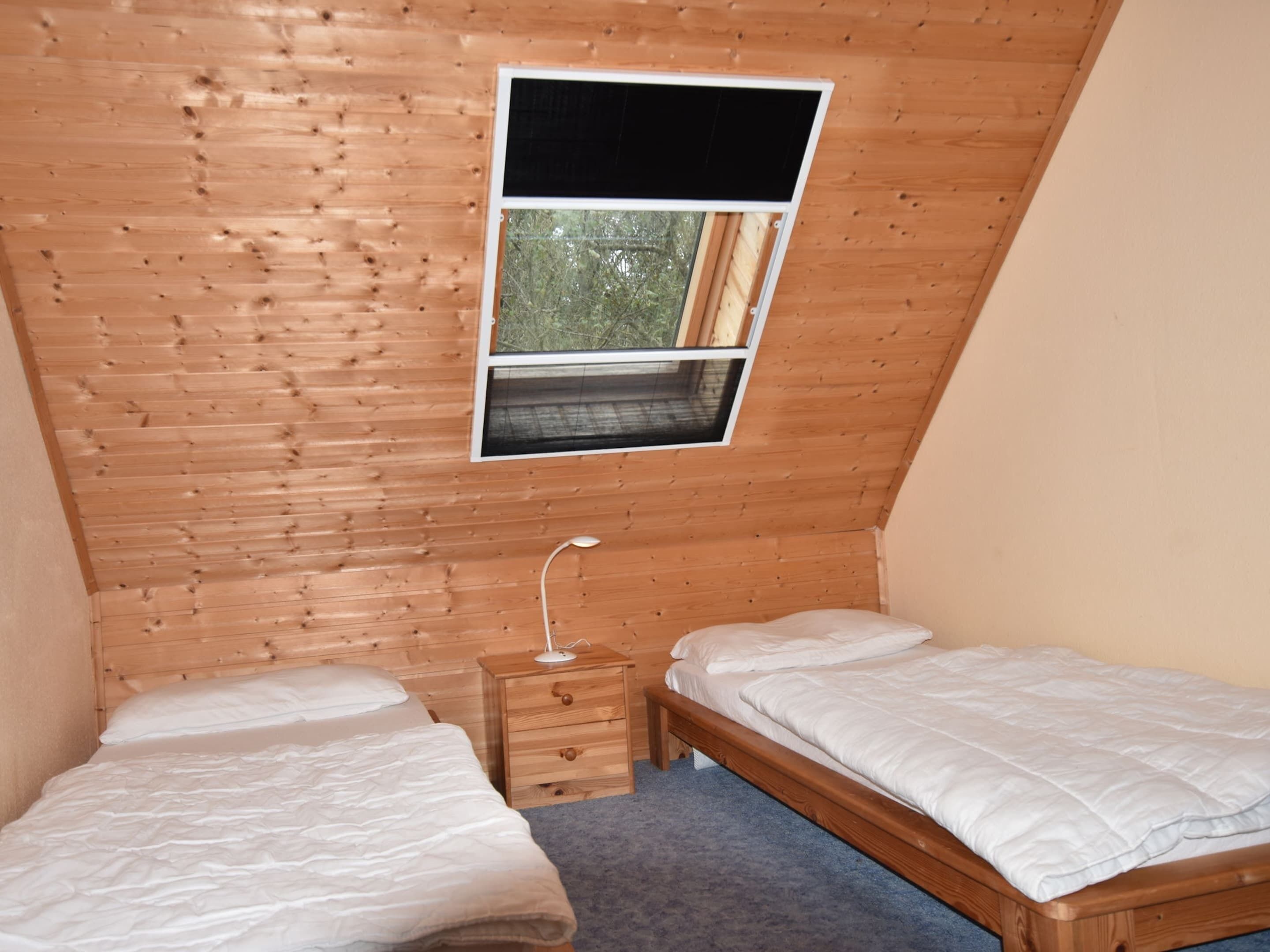 A small, wooden-paneled room with two single beds, a nightstand, and a window with a view of greenery.