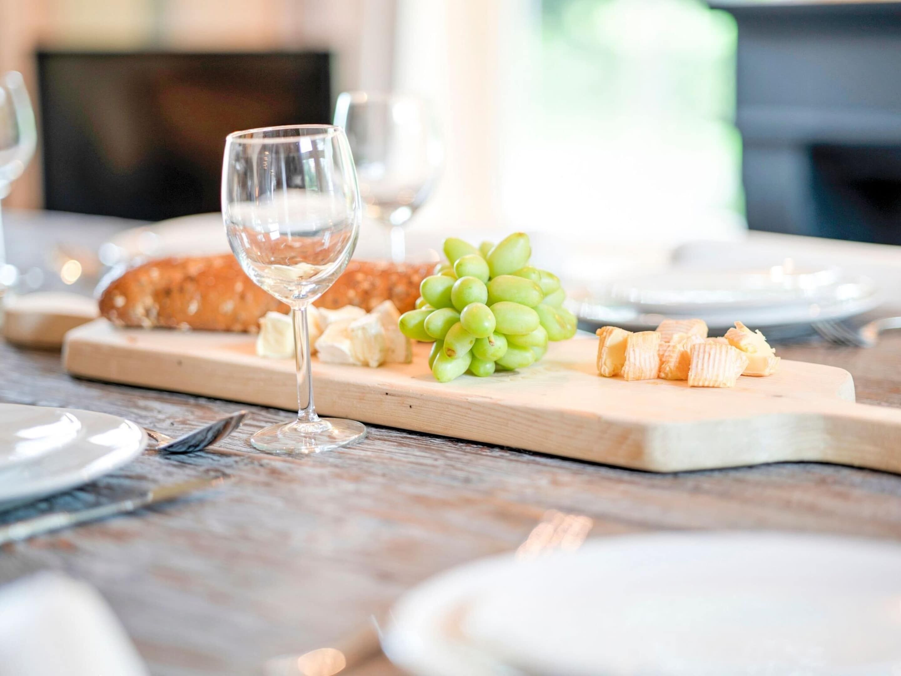 A wooden table with a glass wine glass, a loaf of bread, a bunch of green grapes, and cheese on a wooden board.