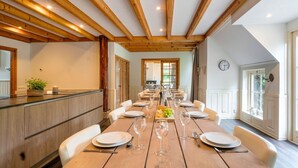 A dining area with a long wooden table set for a meal, wooden beams on the ceiling, and a view of the kitchen through an open door. - Spacious Farmhouse With Sauna, Hottub (Noardburgum)