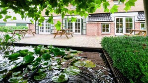A courtyard with a brick building, wooden picnic tables, and a pond with lily pads. - Spacious Farmhouse With Sauna, Hottub (Noardburgum)