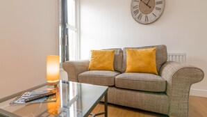 A living room with a sofa, a glass coffee table, a lamp, and a wall clock. - Platform Fishing Quarter Apartments (Hull)