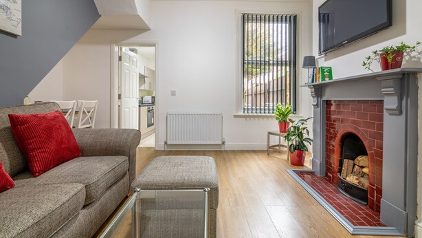 A modern living room with a grey sofa, a red cushion, a glass coffee table, a fireplace with a brick surround, a mounted TV, and a window with blinds. - Platform Fishing Quarter Apartments (Hull)