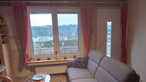 A living room with a sofa, a glass coffee table, a red rug, and a view of a forest through a window with red curtains. - Cozy Holiday Home in Boevange-clervaux With Garden (Wincrange)