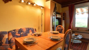 A dining area with a wooden table set for two, a sofa with a colorful pattern, a floor lamp, and a view of the outdoors through a window with curtains. - Cozy Holiday Home in Boevange-clervaux With Garden (Wincrange)