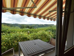 A covered patio with a table and chairs, overlooking a lush garden with yellow flowers and a view of green hills in the distance. - Cozy Holiday Home in Boevange-clervaux With Garden (Wincrange)