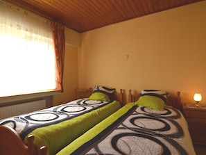 A bedroom with two single beds, wooden headboards, a nightstand with a lamp, and a window with curtains. - Cozy Holiday Home in Boevange-clervaux With Garden (Wincrange)