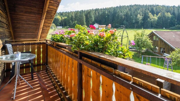 A wooden balcony with a table and chairs, overlooking a garden with pink flowers and a view of a forested area. - Bauernhof in Spandlberg am Hohe Bogen (Neukirchen Beim Heiligen Blut)