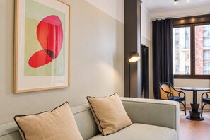 A modern living room with a sofa, a framed abstract painting, a floor lamp, and a view of the city through large windows. - Sweett | Sepulveda XI (Barcelona)