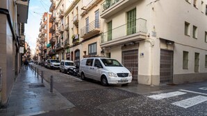 A narrow urban street lined with multi-story buildings, parked cars, and a pedestrian walkway. - Sweett | Verdi Baixos (Barcelona)