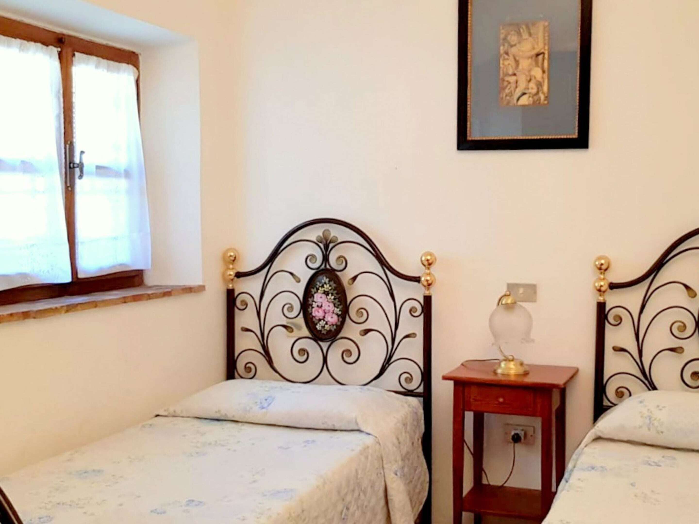 A bedroom with two single beds, a small bedside table with a lamp, a window with curtains, and a framed picture on the wall.