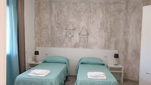 A room with two beds, each with a teal bedspread and white towels folded on top. There are bedside tables with lamps, a wall-mounted decoration, and a plain wall with a textured finish. - Caletta sul Mare (Siniscola)