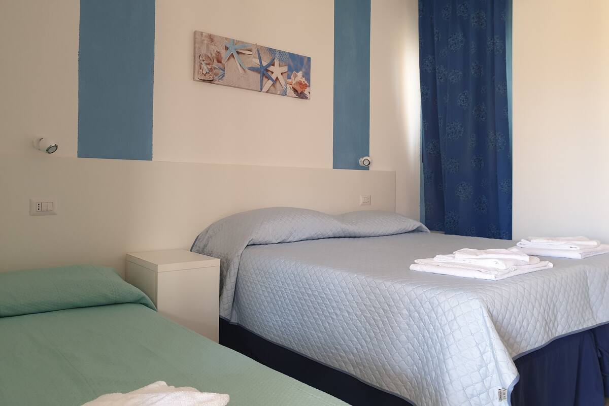 A room with two beds, a blue and white color scheme, a painting on the wall, and a blue curtain.