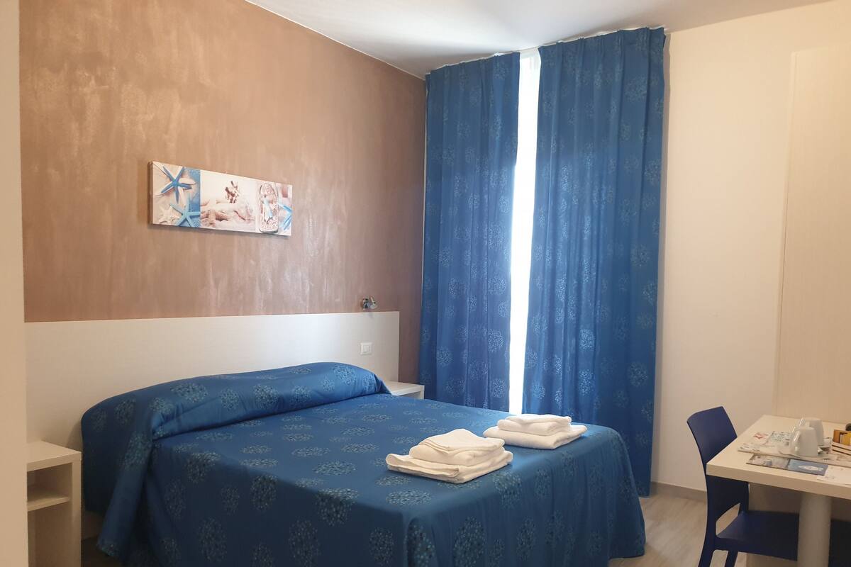 A hotel room with a bed, a desk, a chair, a window with curtains, and a wall-mounted artwork.