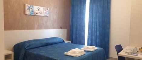 A hotel room with a bed, a desk, a chair, a window with curtains, and a wall-mounted artwork.