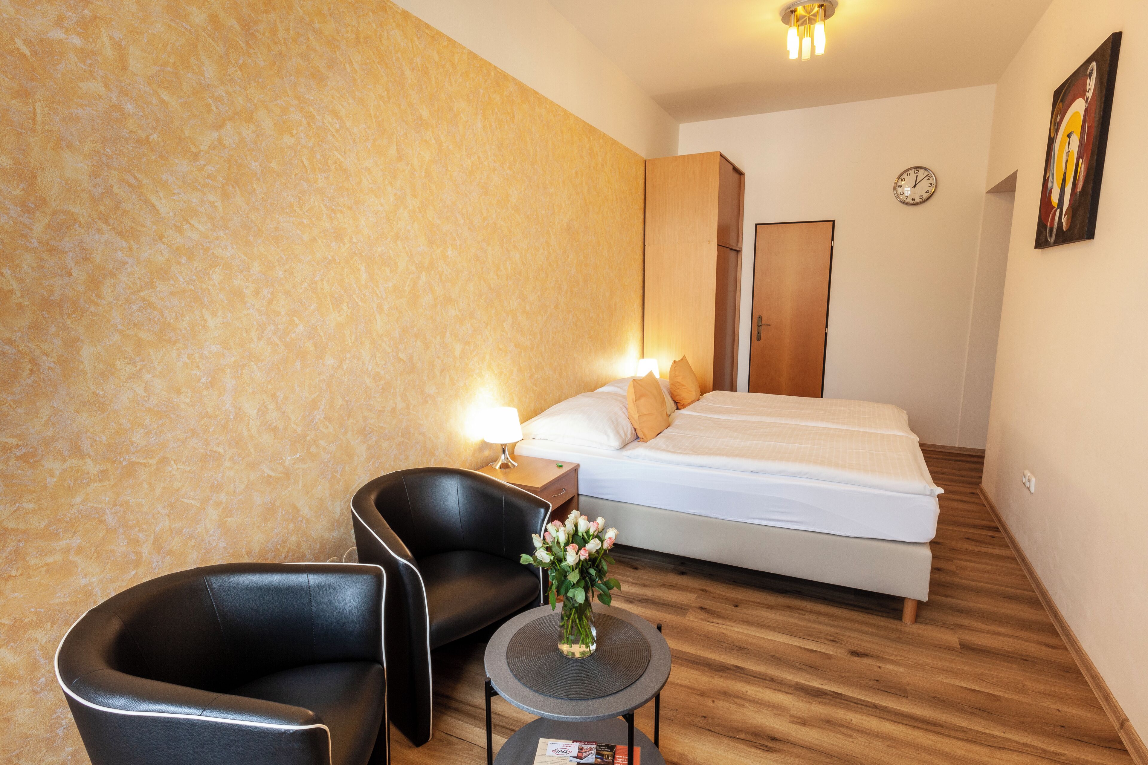 A hotel room with a bed, a chair, a small table with a vase, a nightstand, and a wall clock.