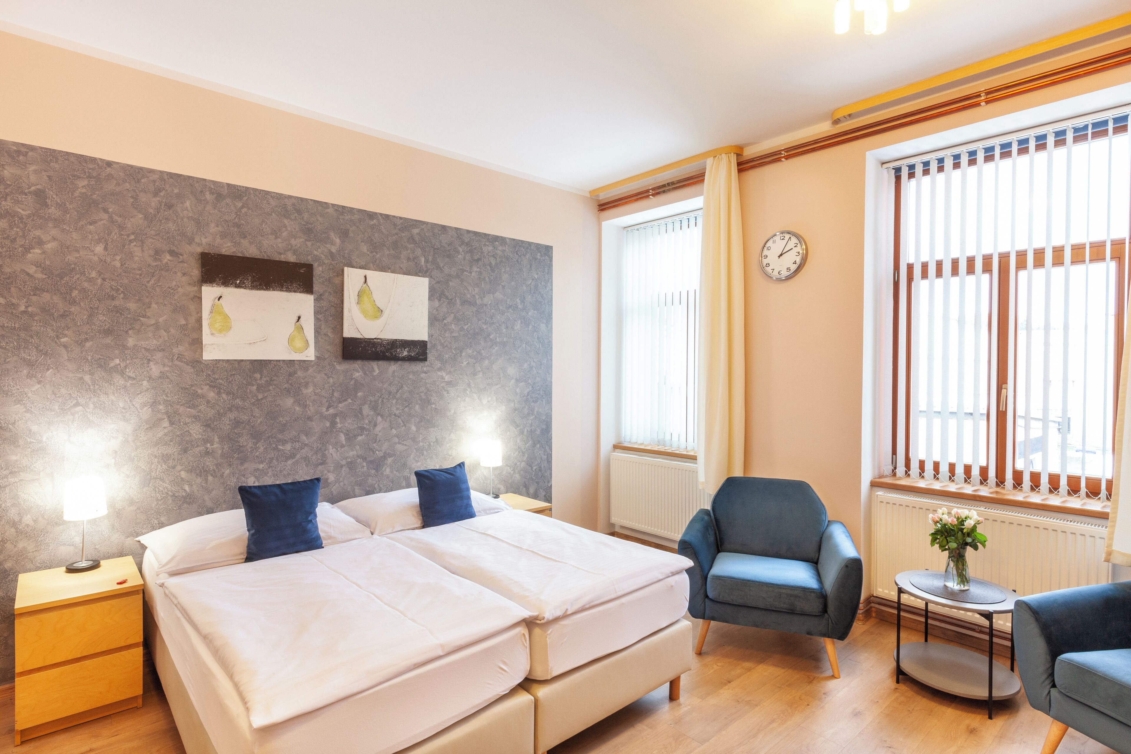 Ein Hotelzimmer mit einem Doppelbett, zwei Nachttischen, einem Sessel, einem kleinen Tisch mit einer Vase und einem Fenster mit Jalousien.
