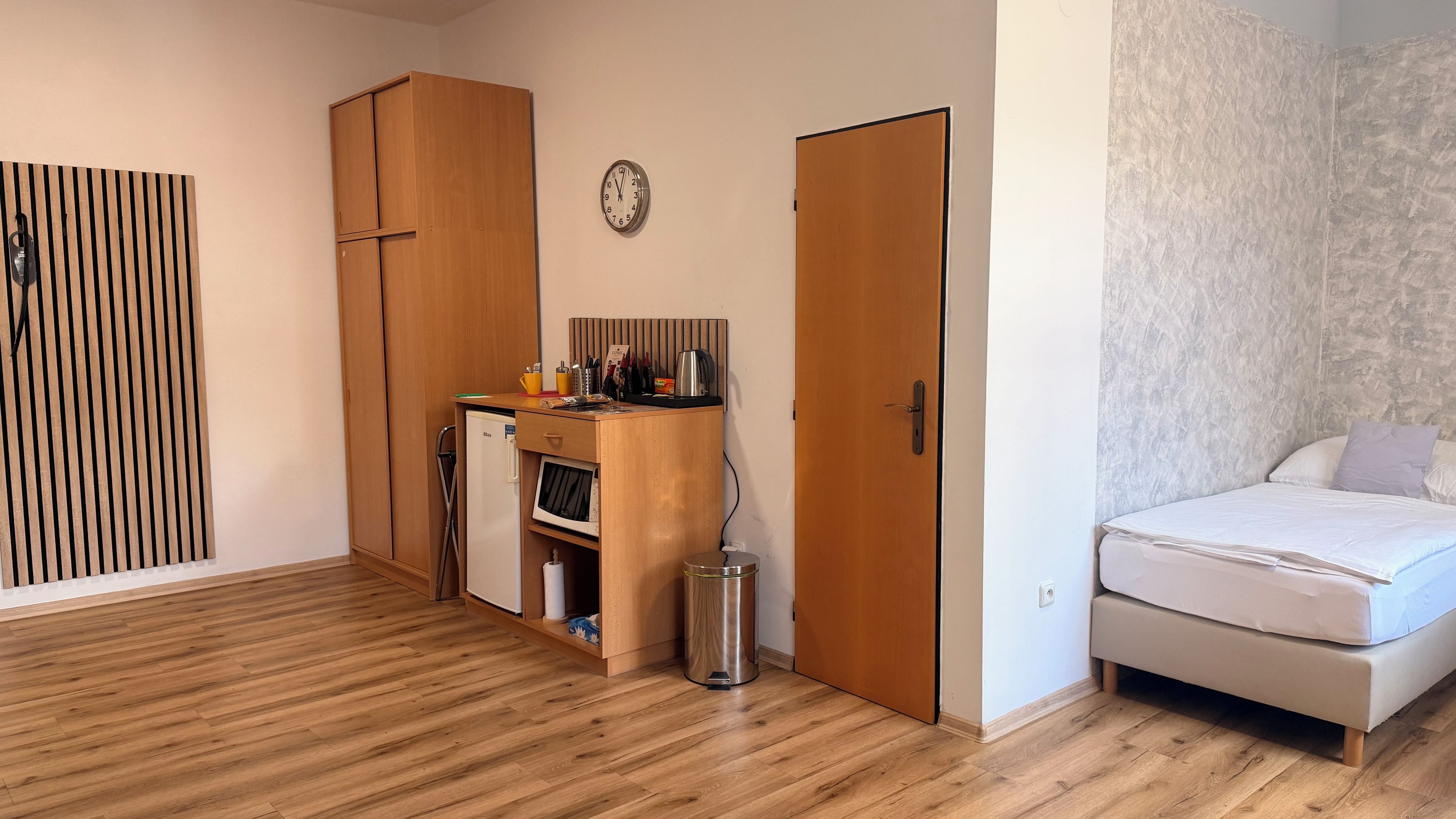 Un dormitorio con cama, armario, pequeña kitchenette y un bote de basura.